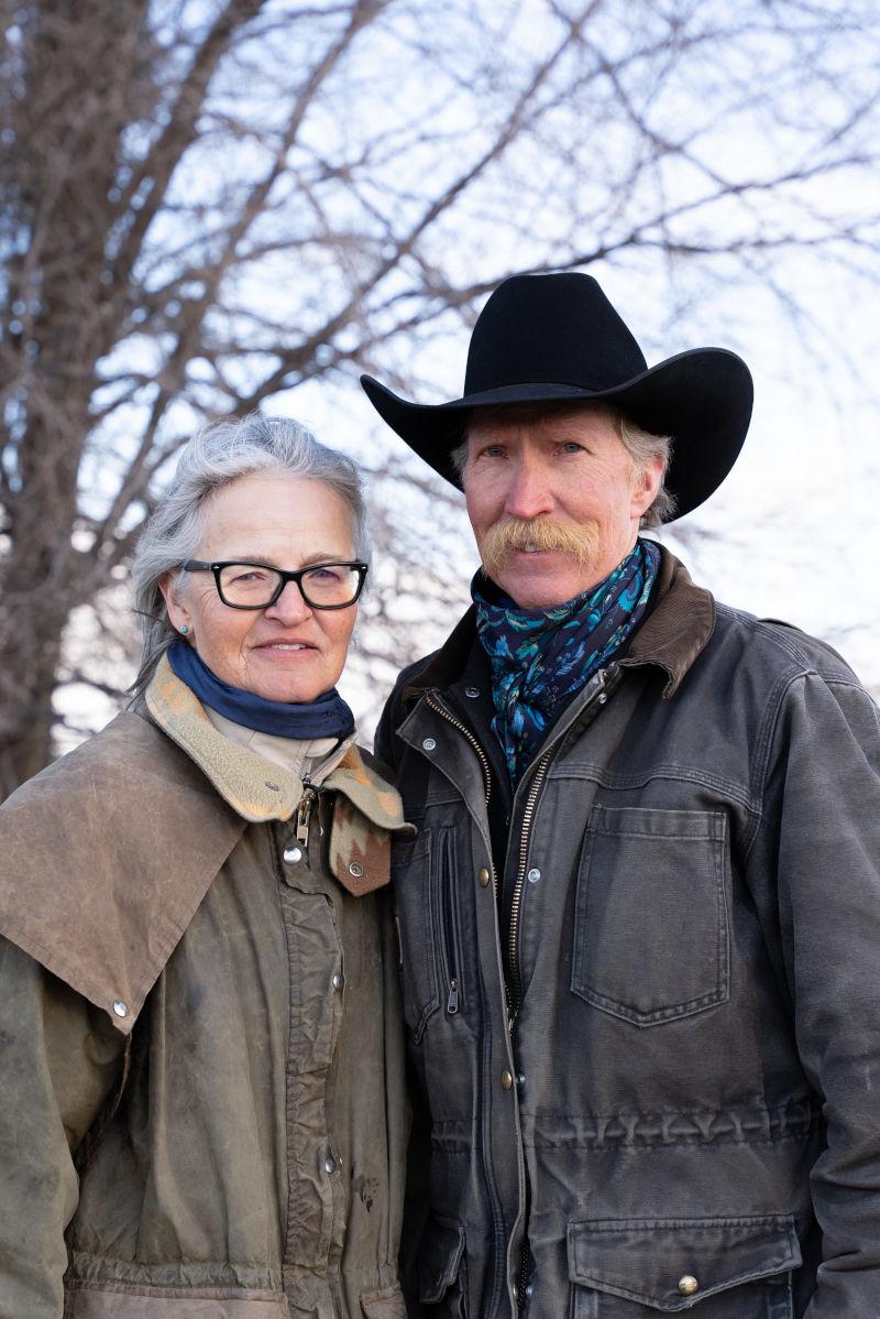 一名戴着西部帽子、留着牛仔小胡子的男子站在田野里，旁边是一名身穿棉衣式牧场大衣的女子.